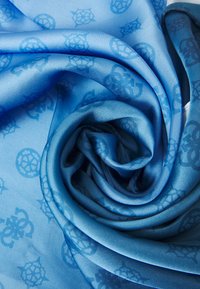 Écharpe en soie bleue avec une texture lisse, présentant un motif subtil de designs nautiques superposés. Roulée, mettant en valeur une profondeur de couleur vibrante.