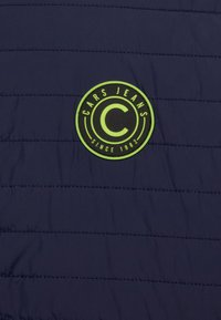 Marinblå quiltad tyg med en rund svart och neongrön logotyp som har texten "CARS JEANS" och "SINCE 1982".