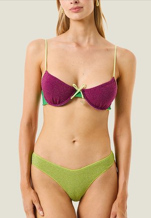 Donna che indossa un bikini glitter bicolore con coppe viola, fascia inferiore verde e slip del bikini verde lime su sfondo uniforme.