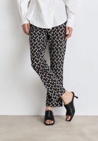 Black and cream patterned trousers with a straight cut, paired with open-toed black mules featuring a low heel. White button-up shirt worn.
