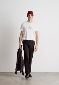 Camiseta branca com um logo circular, calças escuras, sapatos pretos e um gorro bordô; segurando um suéter escuro; fundo minimalista.