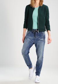 Prošívaná tmavozelená bomber bunda, světle modrý žebrovaný svetr a vybledlé modré jogger džíny. Bílí tenisky dotvářejí celý outfit.