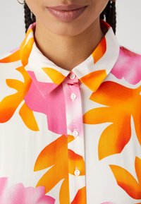 Blusa floreale con sfondo bianco decorato da grandi motivi a foglia arancioni e rosa, rifinita con colletto e chiusura con bottoni.