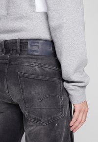 Felpa grigia indossata con jeans di denim grigio scuro che presentano un'etichetta in pelle G-Star RAW, cuciture sottili e tasche posteriori angolate.