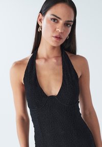 Svart halterneckklänning med ett texturerat mönster. Djup V-ringning och figurnära design. Guldörhängen i ringform syns. Ljus bakgrund.