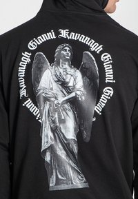 Sudadera negra con un prominente gráfico de un ángel plateado en la parte posterior, con texto en letras blancas en negrita que rodea el diseño.