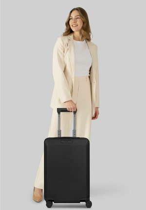 Jonge vrouw in beige pak met een zwarte koffer op wieltjes met uitgetrokken handvat, staand tegen een effen grijze achtergrond.