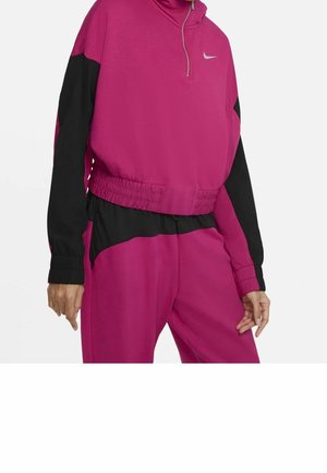 Kvinde iført en magenta og sort Nike træningsdragt med en lynlåstop og elastisk talje, stående mod en hvid baggrund.