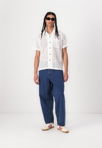 White short-sleeve shirt with cut-out patterns, button closure, paired with loose blue denim jeans and beige shoes with red stripes.