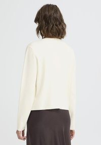 Kremno obarvan skrajšan pulover iz mehke tkanine, ki ima dolge rokave in okrogel izrez, v kombinaciji s temno rjavim krilom.