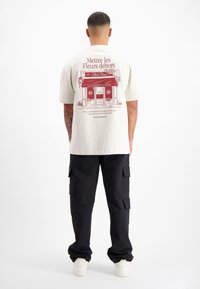 Beige t-shirt met korte mouwen, voorzien van een rode illustratie van een bloemenwinkel en tekst op de achterkant, gecombineerd met zwarte cargobroeken en witte sneakers.