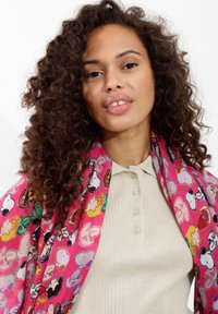 Écharpe rose avec des motifs de dessins animés, drapée autour d'une personne, portant un haut beige à côtes en dessous et des cheveux bouclés encadrant le visage.