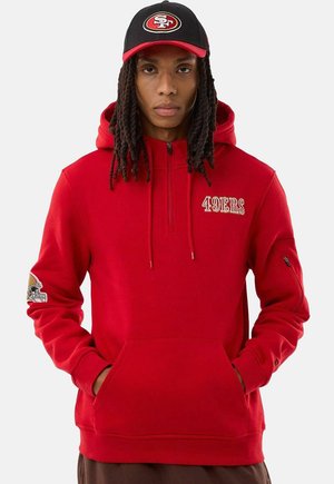 Persona che indossa una felpa con cappuccio rossa dei San Francisco 49ers e un cappello nero dei 49ers con le mani nella tasca della felpa su uno sfondo semplice.
