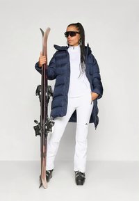 Γυναίκα με άσπρη στολή σκι, ναυτική τζάκετ από πούπουλα, μαύρες μπότες σκι και γυαλιά ηλίου, κρατώντας ένα ζευγάρι σκι και στέκεται μπροστά σε απλό φόντο.