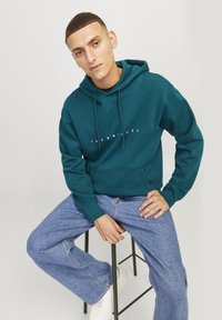 Teal hettegenser med snøring, frontlomme og hvit logo tekst. Brukt med lyseblå jeans og hvite joggesko, sittende på en krakk.