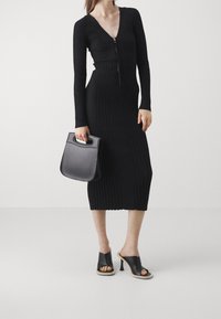 Robe noire en maille côtelée avec manches longues et fermeture éclair à l'avant. Associée à un sac à main noir et des sandales noires à talons ouverts.