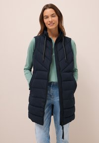 Piumino blu navy senza maniche con colletto alto, zip e lacci. Presenta un design trapuntato con tasche frontali, indossato sopra un top verde e jeans.