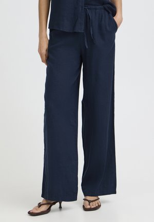 Femme portant un pantalon large bleu marine à taille élastique avec cordon de serrage et des sandales à talons noires, debout devant un fond uni.