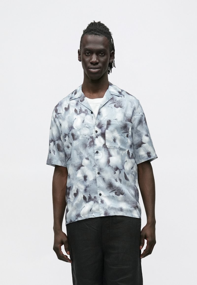 Hombre con cabello trenzado, vistiendo una camisa de manga corta con botones de patrón azul y blanco sobre una camiseta blanca, y pantalones negros, de pie.