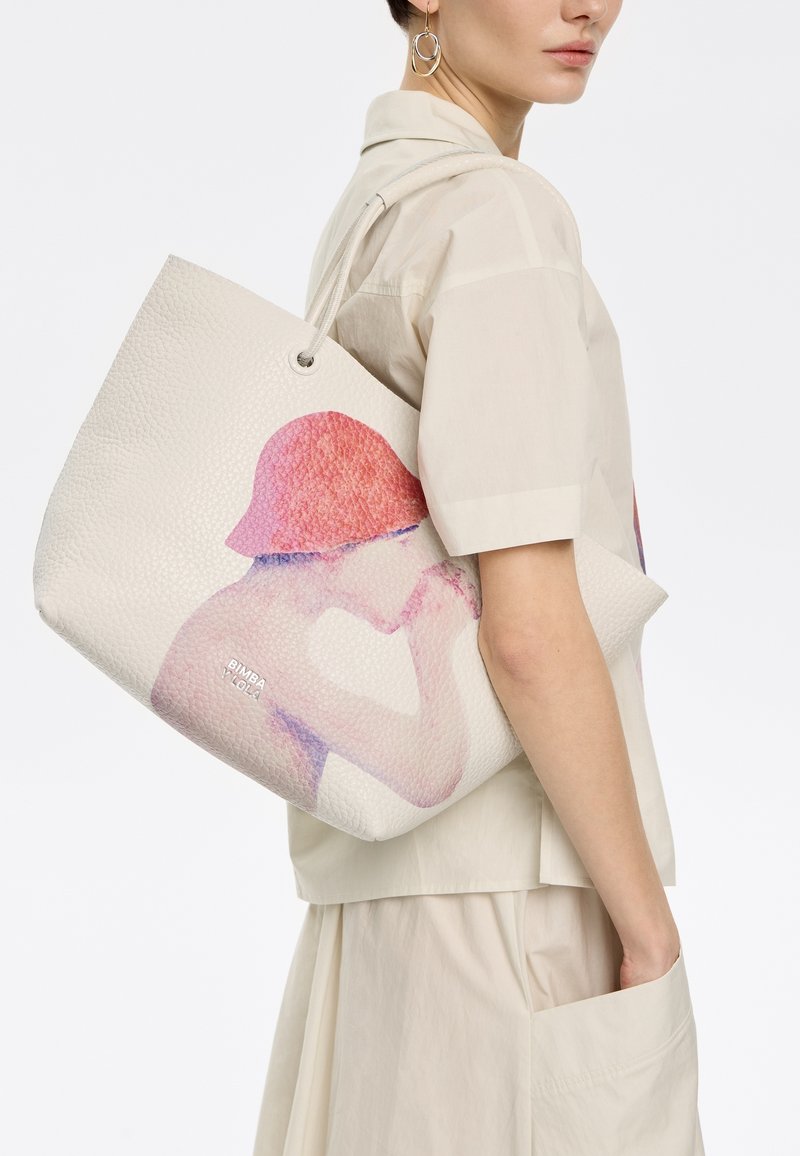 Witte tote bag met textuur, voorzien van een levendig roze en blauwe grafisch ontwerp aan één zijde. De tas heeft een ronde handgreep en een logo aan de voorkant.