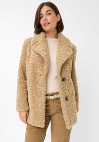 Tan, donzige textuur jas met een brede revers, dubbele knoopsluiting en zijzakken. Draag over een beige top en bruine broek.