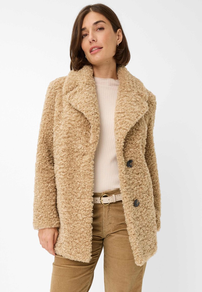 Cappotto di colore tan, con una consistenza morbida, colletto ampio, chiusura a doppi petti con bottoni e tasche laterali. Indossato sopra una maglietta beige e pantaloni marroni.