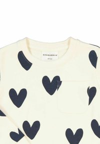 Krämfärgad långärmad tröja med marinblå hjärtmönster och en liten ficka på vänster sida. Mjuk tyg med rund halsringning.