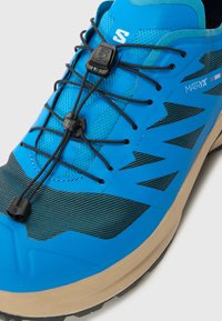 Chaussure de randonnée bleue avec un dessus texturé, des accents en maille noire, et un système de laçage rapide, avec une semelle beige et un crantage en caoutchouc.