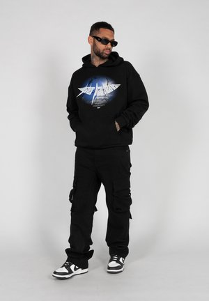 Black hoodie with a graphic design, black cargo pants, and black and white sneakers. Model poses with hands in pockets against a neutral background.