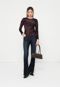 Lange mouwen shirt met een paars en zwart abstract patroon, gecombineerd met donkere flared jeans, een kleine grijze handtas vasthoudend, en zwarte schoenen.