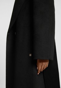 Black wool coat featuring a tailored design, textured fabric, and gold "HUGO" logo accent on the sleeve. Double-breasted with buttons.