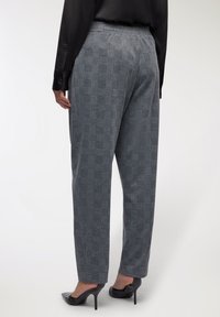 Kobieta ubrana w szare w kratkę spodnie o prostym kroju i czarne, spiczaste szpilki, stoi tyłem do aparatu na białym tle.