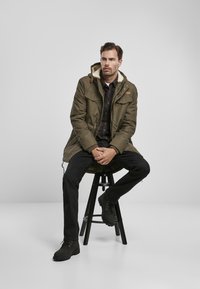 Parka verde măsliniu cu glugă căptușită cu blană fake, asortată cu o cămașă cu carouri închise, blugi negri și botine negre, așezat pe un taburet.