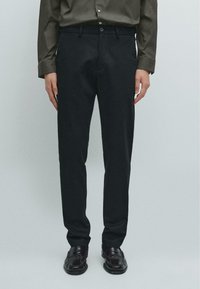 Černé kalhoty na míru se slim fit, vyrobené z měkké tkaniny, s klasickým pasem a zapínáním na knoflík. Nosené s tmavými botami.