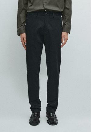 Pantaloni neri su misura con vestibilità slim, realizzati in tessuto morbido, dotati di vita standard e chiusura con bottone. Indossati con scarpe scure.