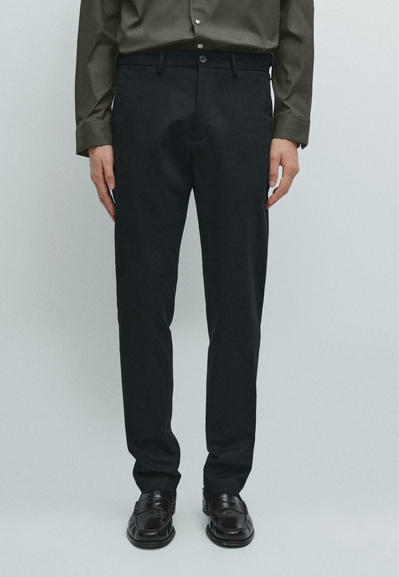 Černé kalhoty na míru se slim fit, vyrobené z měkké tkaniny, s klasickým pasem a zapínáním na knoflík. Nosené s tmavými botami.