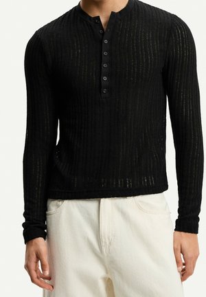 Man draagt een zwart geribd Henley-shirt met lange mouwen en knopen en off-white broek tegen een effen achtergrond.