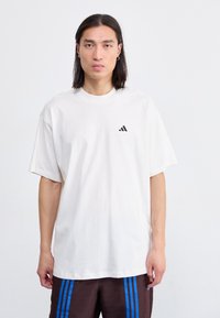 Balts pārmērīgs T-krekls no kokvilnas, ar melnu Adidas logotipu kreisajā krūtī. Gluda tekstūra, apaļa kakla izgriezums un īsas piedurknes.
