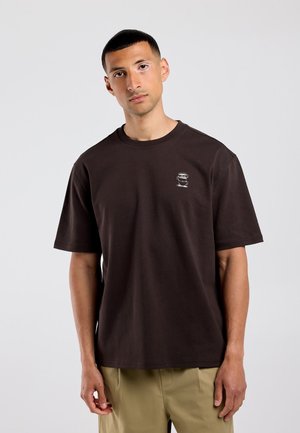 Jonge man met kort haar, gekleed in een donkerbruin t-shirt met een klein wit patroon en licht beige broek tegen een effen grijze achtergrond.