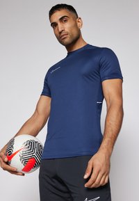 Mørkeblå atletisk skjorte med en tætsiddende pasform, korte ærmer og et hvidt Nike-logo; modellen holder en sort og hvid fodbold.