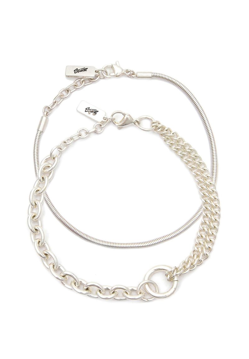 Dvoje prepletených strieborných náramkov s karabínkovými zapínaniami a malými obdĺžnikovými visačkami s vyrytým podpisom.