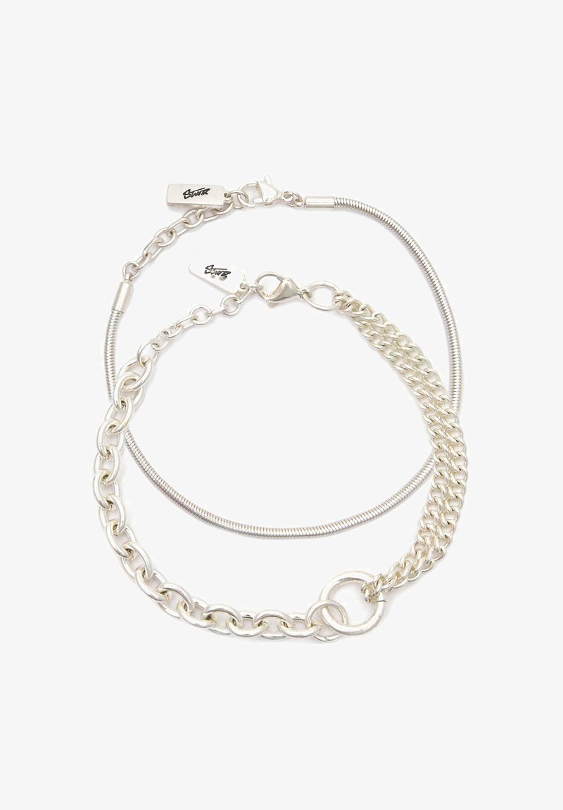 Dvoje prepletených strieborných náramkov s karabínkovými zapínaniami a malými obdĺžnikovými visačkami s vyrytým podpisom.