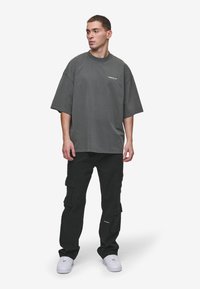 Szürke, oversized póló, fekete cargo nadrággal párosítva. A pólón kicsi logó található a mellen; a nadrágon több oldalsó zseb van. Fehér sportcipő.