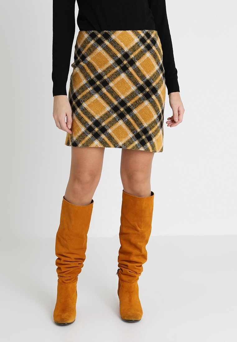 Femme portant une mini-jupe à carreaux jaune et noire, un haut noir à manches longues, et des bottes hautes moutarde lâches, debout sur un sol blanc.