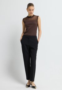 Bruine geribbelde mouwloze top met doorschijnende ruches op de schouders, gecombineerd met zwarte getailleerde broeken en zwarte ballerina's met puntige neuzen en gouden accenten.