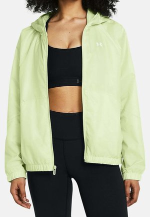 Vrouw draagt een lichtgroen, lichtgewicht jack met rits over een zwarte sport-bh en zwarte high-waist leggings, staand tegen een witte achtergrond.