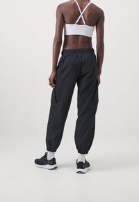 Kobieta w białym sportowym staniku, czarnych spodniach typu cargo, białych skarpetkach i czarnych butach sportowych, stojąca plecami do kamery.