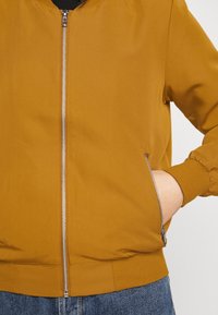 Kurtka bombowiec w musztardowym kolorze z zamek błyskawicznym z przodu, ryflowanymi mankietami i pasem. Posiada boczne kieszenie oraz gładką, matową fakturę.
