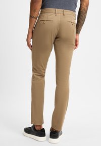 Beige Chinos mit schmaler Passform, ausgestattet mit zwei Gesäßtaschen mit Knöpfen und einer glatten Textur, kombiniert mit schwarzen Sneakers und weißen Sohlen.