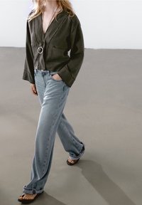 Femme portant une veste olive ample, un jean large bleu clair et des tongs noires, marchant sur un sol gris lisse à l’intérieur.